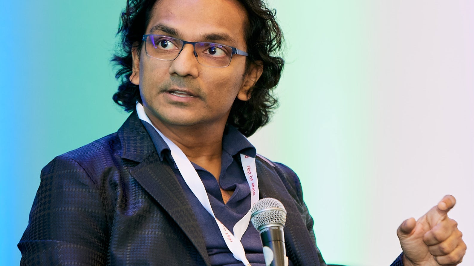 a photograph of a man seated, wearing a blazer and speaking into a microphone.