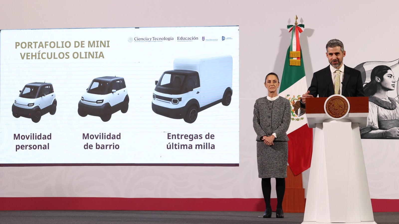A presentation showcasing the 'Portfolio of Mini Vehicles Olinia', featuring three models labeled for personal mobility, neighborhood mobility, and last-mile deliveries. Two presenters stand beside the display; a man at a podium and a woman to the side, with a Mexican flag in the background.