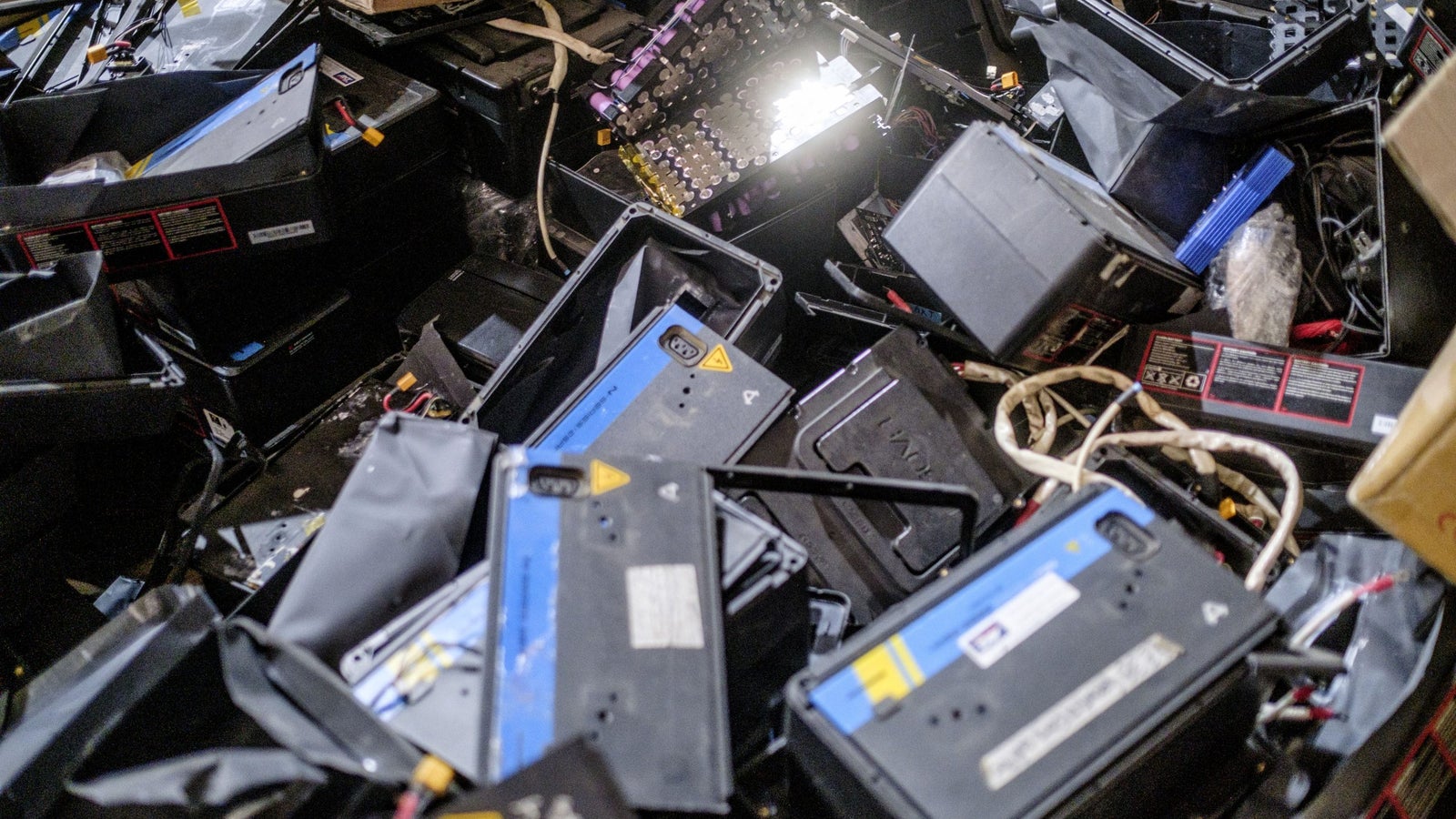 A chaotic pile of discarded batteries and battery casings scattered across a surface, with some cables and a cardboard box partially visible in the background.