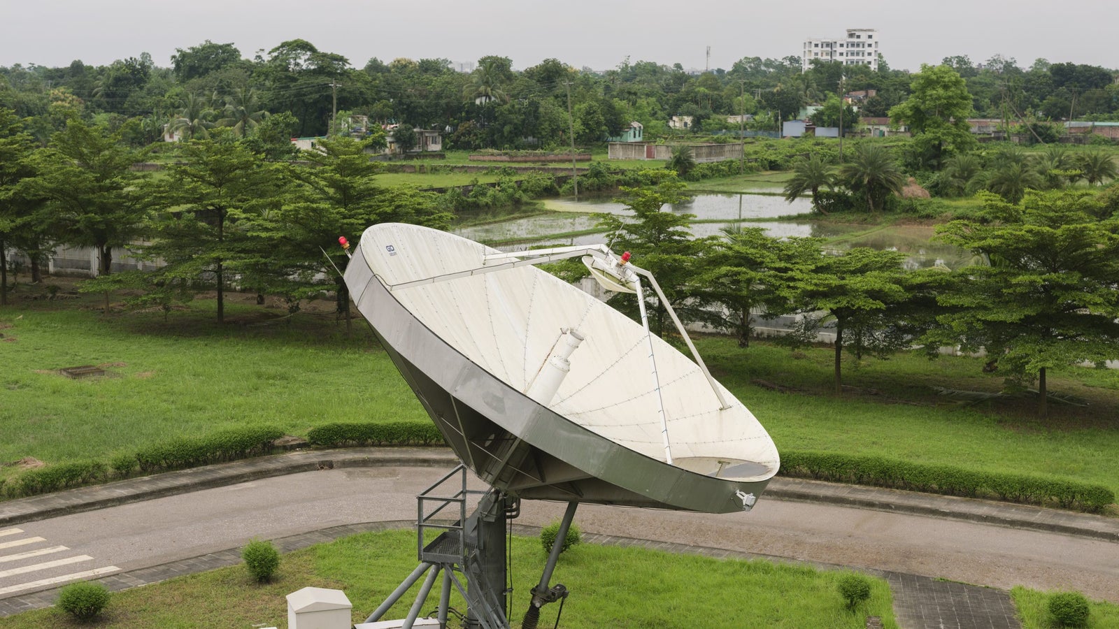 A large satellite dish in a green area, with trees and a pond in the background under a cloudy sky.