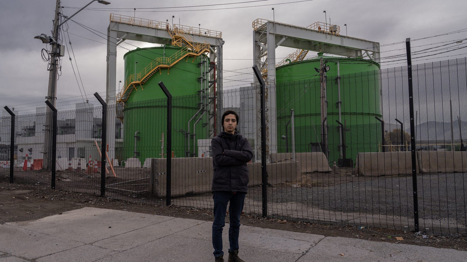 A photograph showing a man, standing with his arms crossed and wearing black clothing, in front of two large green industrial tanks.