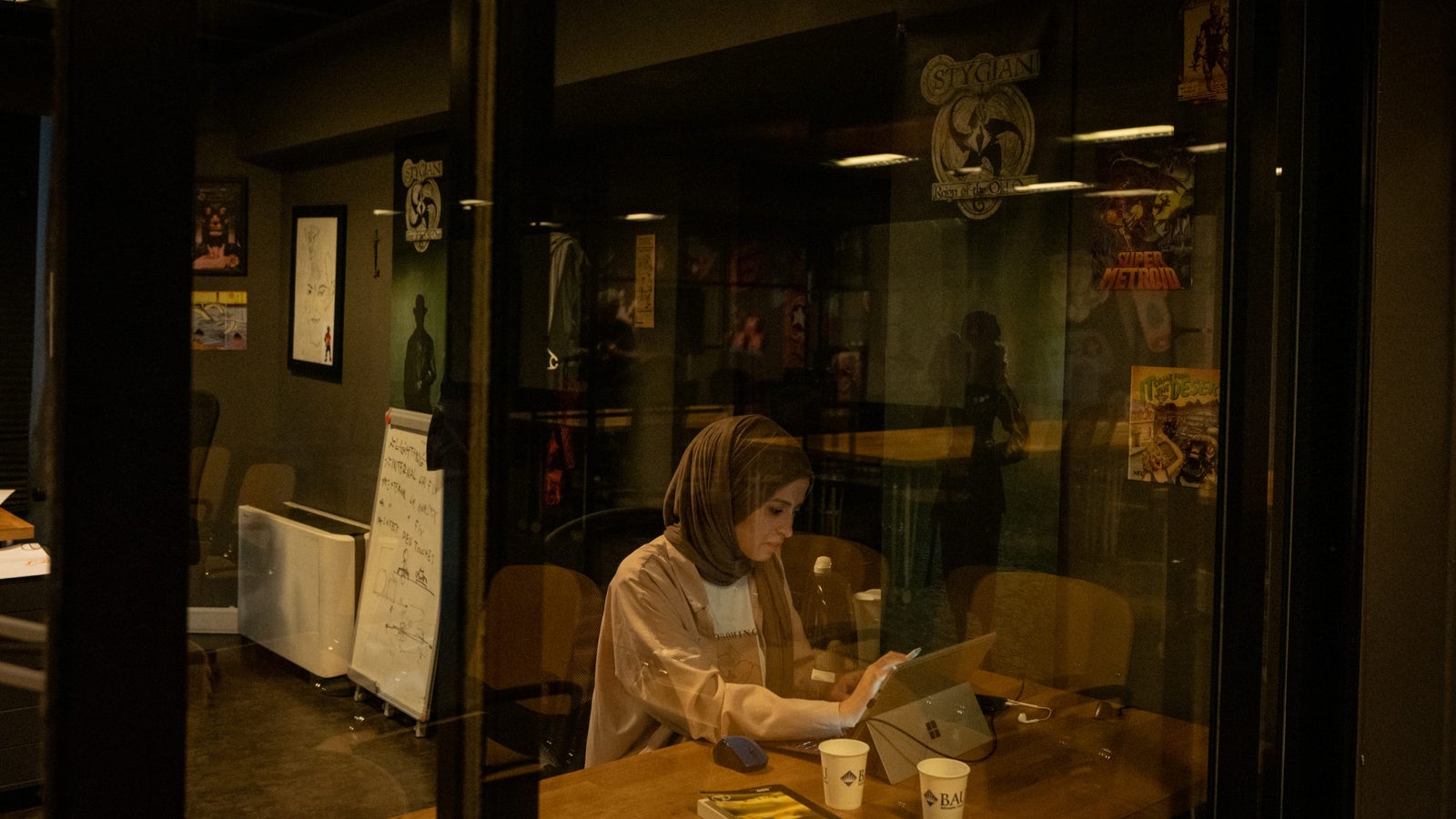 A woman wearing a hijab sits at a wooden table in a dimly lit office space, engaged with a tablet and laptop; behind her, walls adorned with various posters and a whiteboard with handwritten notes are visible.