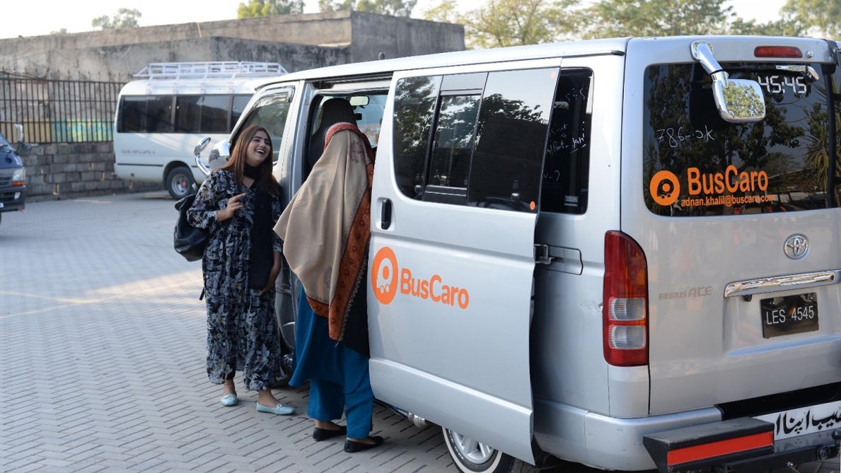 A woman in a patterned outfit laughs while standing next to a van with "BusCaro" branding, as another woman in traditional attire enters the vehicle. The scene is set in a parking area with a few other vehicles in the background.