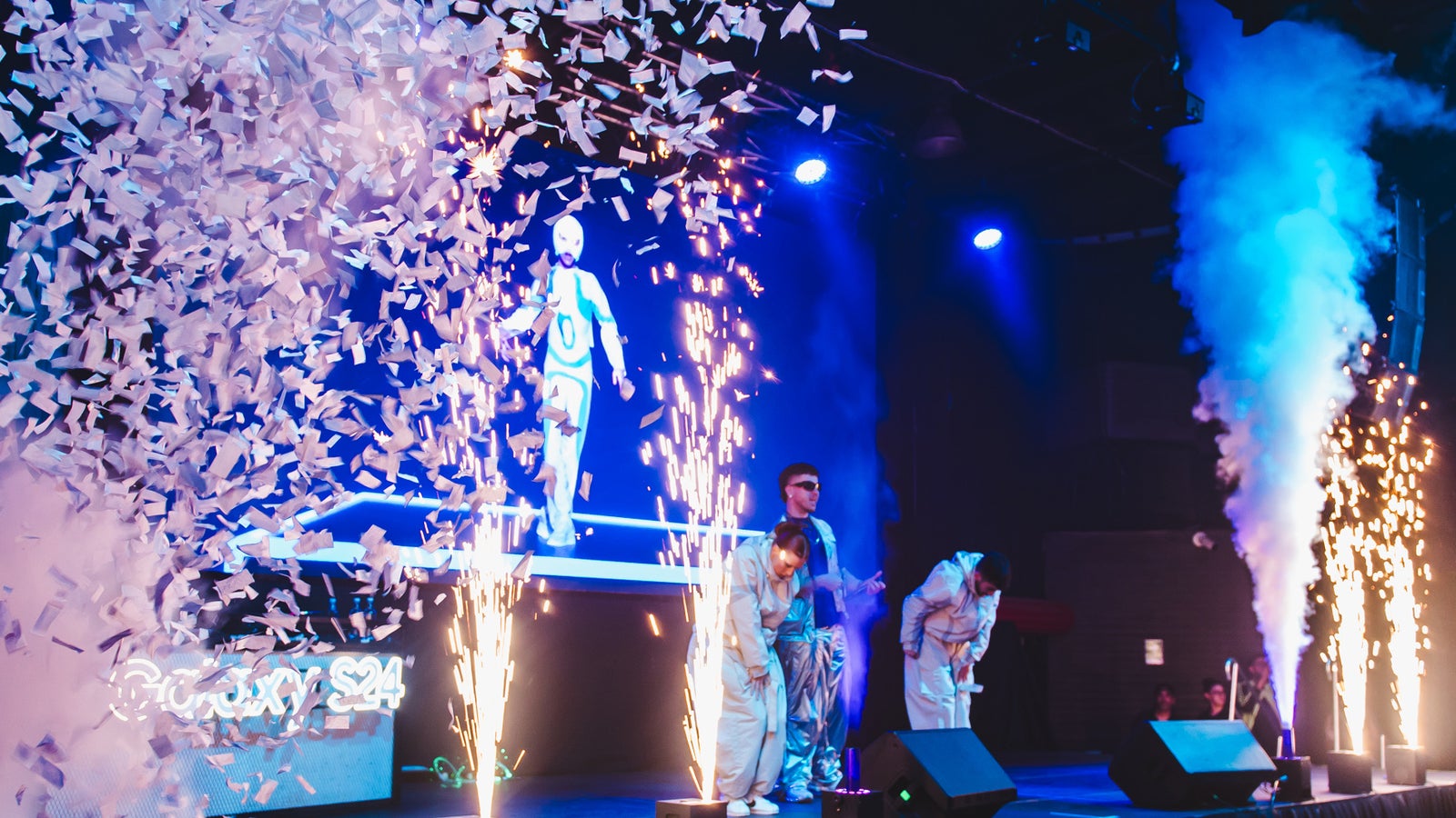 A photograph of a stage with fireworks and confetti, with performers standing behind it lit by blue lights.