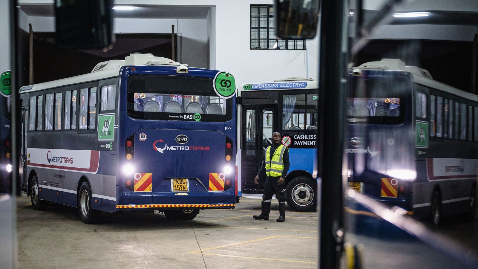 Two buses are parked in a garage with one driver in a yellow reflective vest standing beside them. One bus is labeled "METROTRANS" and the other has "EMBASSAVA ELECTRIC" signage. The environment appears dimly lit, with some reflections visible on the surfaces.