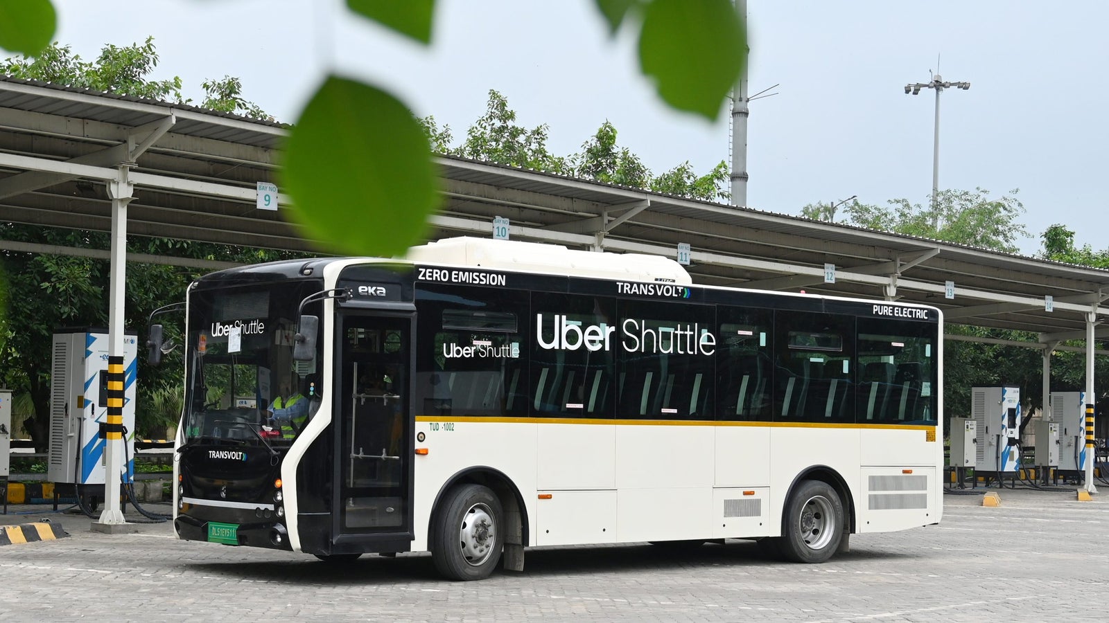 A bus with "Uber Shuttle" branding is parked at a charging station under a roofed canopy, surrounded by greenery.