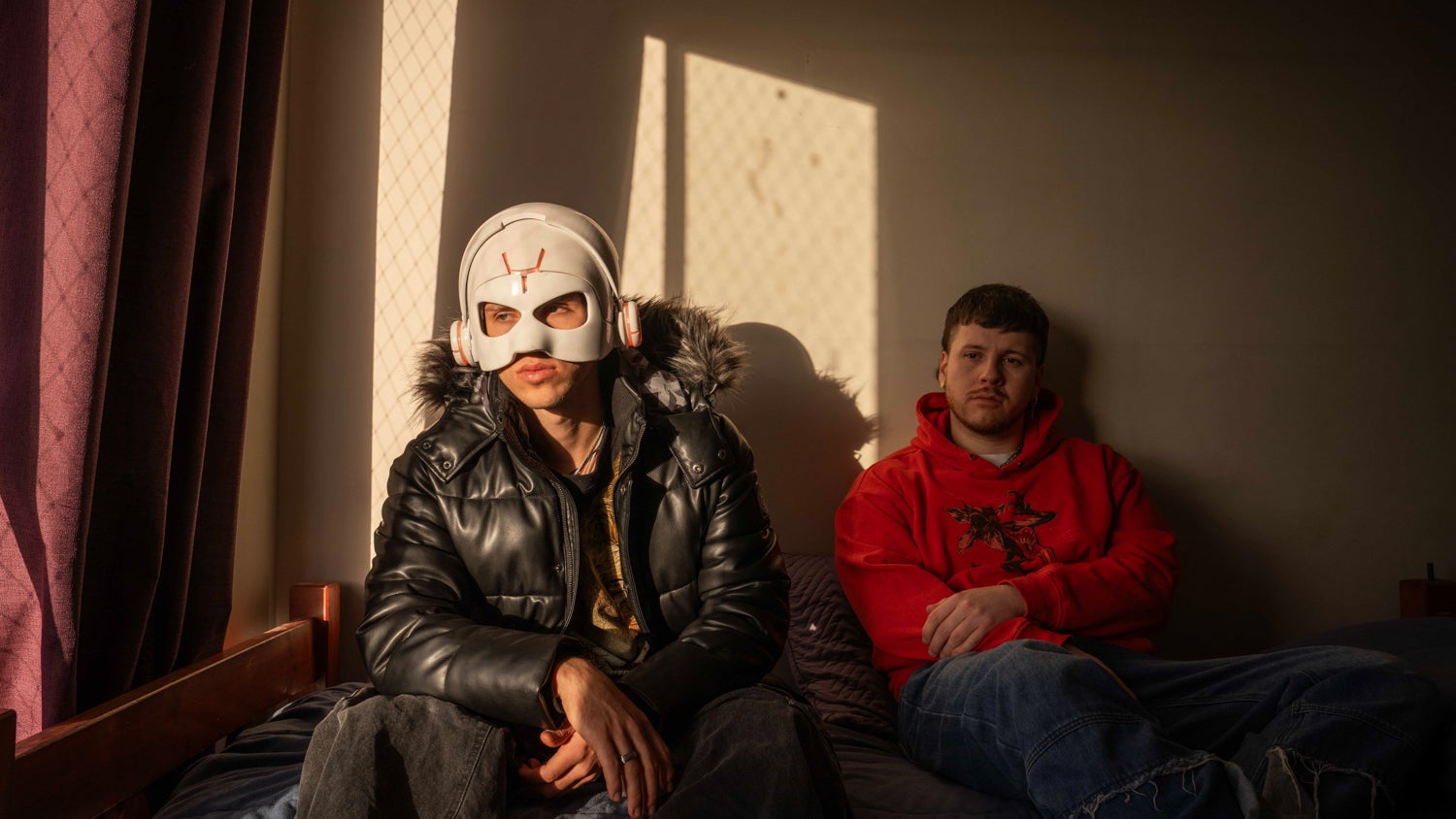 Mauricio Bustos (right) together with his brother Marcelo Bustos (left) who wears a white robotic mask. Both are sitting on the bed in the small room they share in Santiago, Chile, sunlight shining through one side.