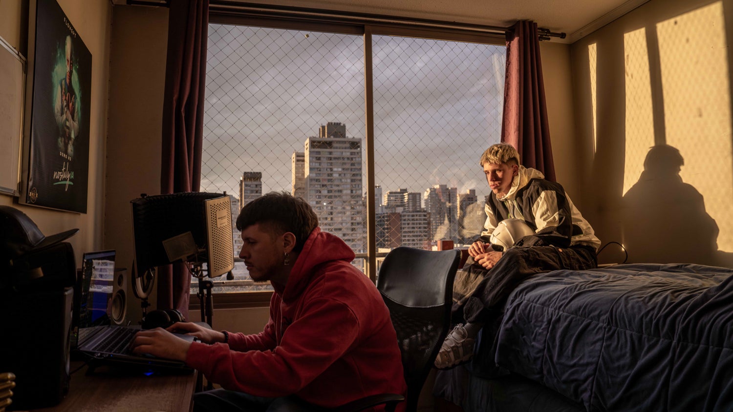 Mauricio Bustos with his brother Marcelo Bustos are in their room. Bustos is working on a laptop while Marcelo sits on the bed watching him. Outside their window are tall buildings and a grey sky.