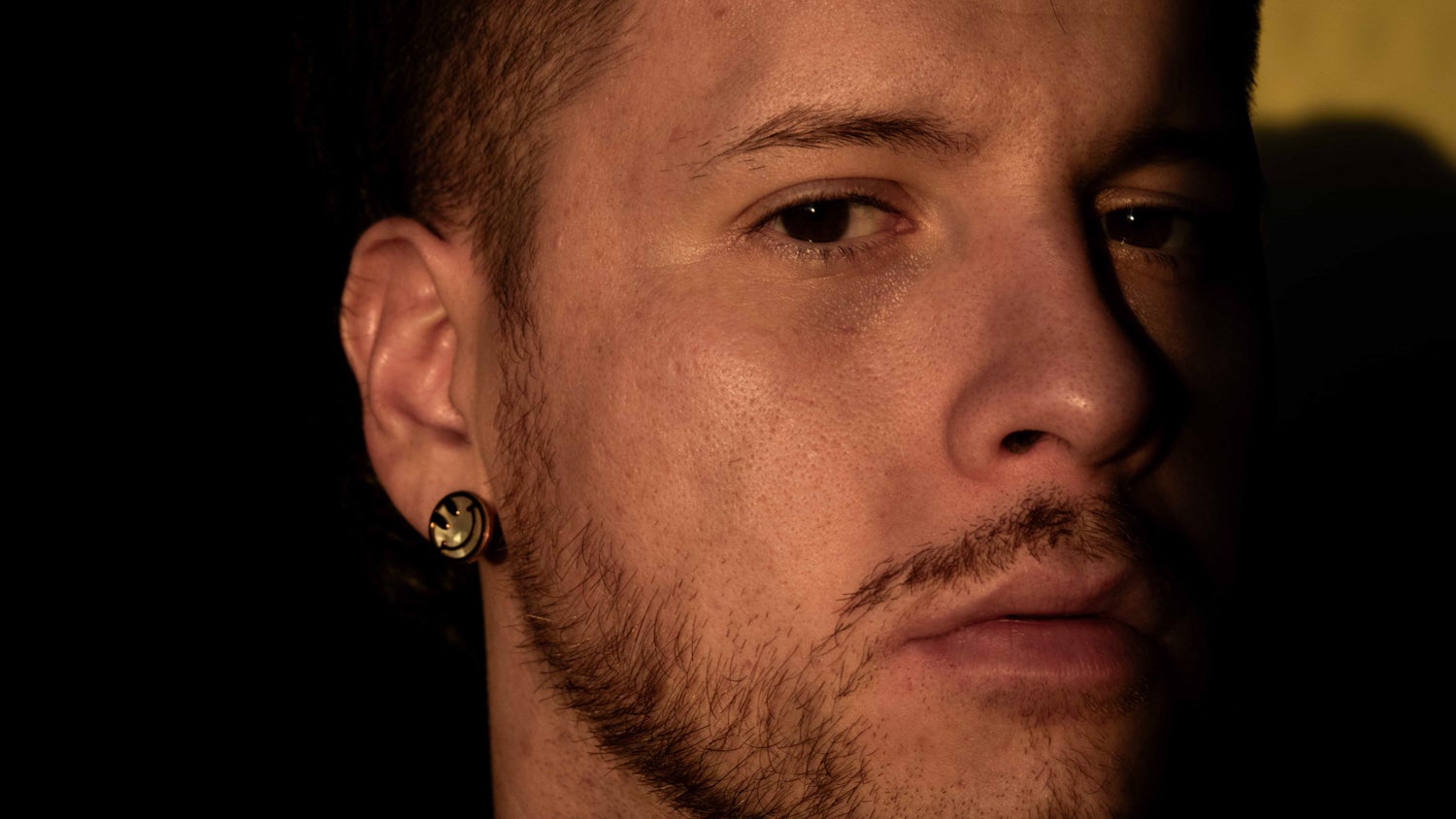 A closeup portrait of Mauricio Bustos, his face partially in shadows, wearing a smiley face earring.