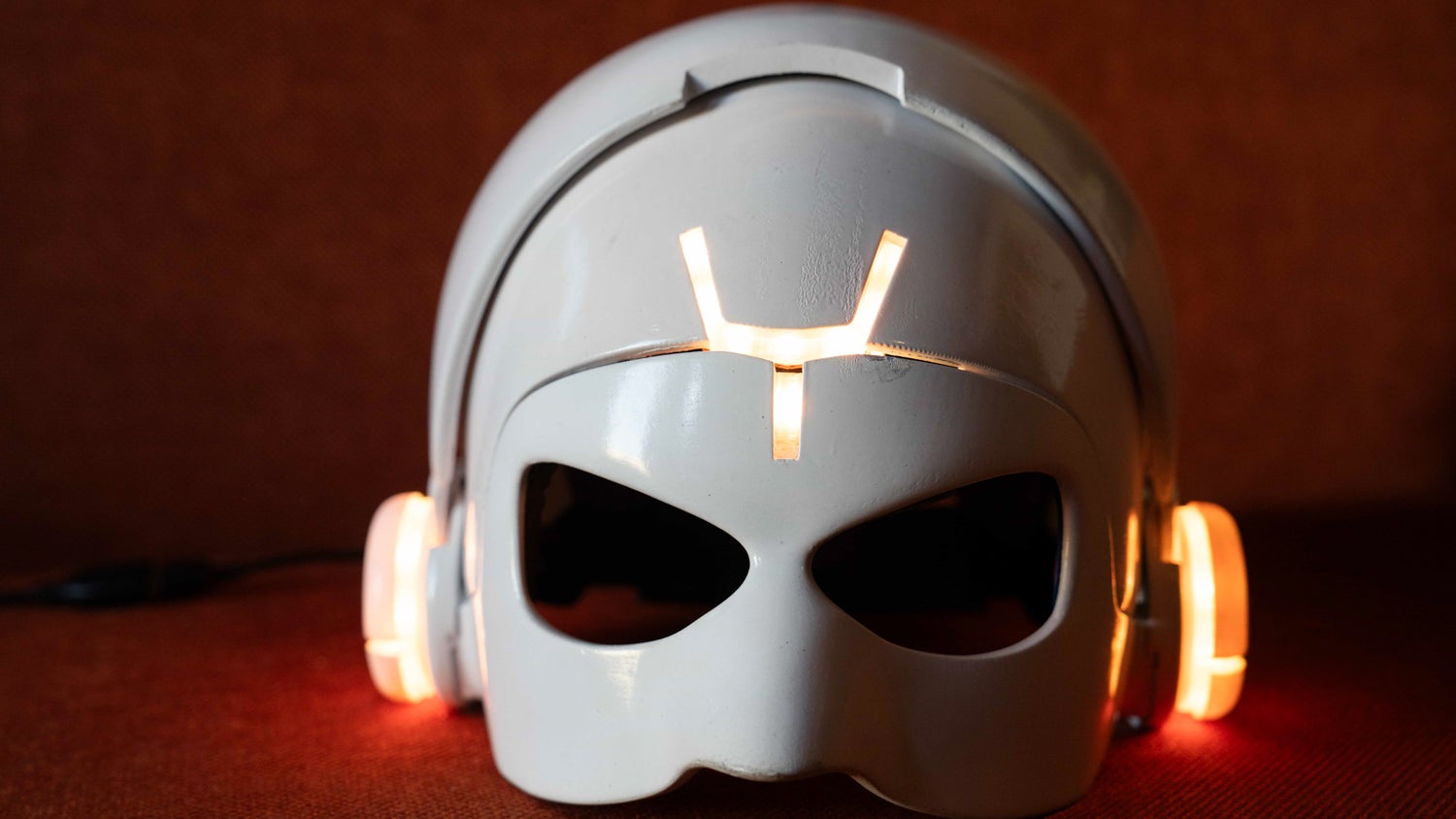 A photograph of a white robotic mask that identifies FlowGPT on an orange cloth surface. The mask wears a pair of headphones which are lit up in an orange-coloured light.