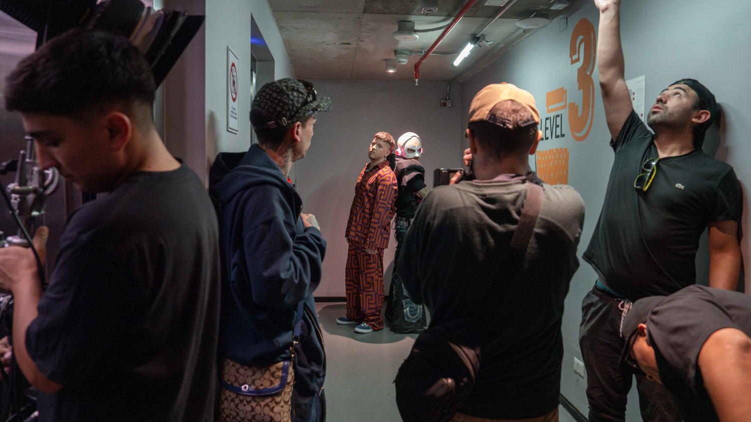 A photograph of a music video recording of FlowGPT in a grey corridor. Mauricio (left) and Marcelo (right, wearing the helmet) are standing with their backs against each other. There are multiple production crew members in the frame.