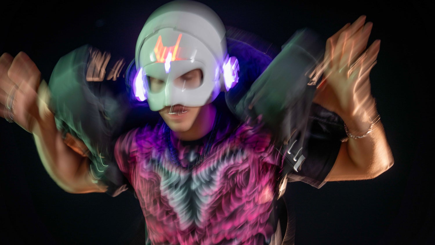 A blurry photo of a man wearing a white robotic helmet and a tshirt with a striking pink design, his jacket and hands are held up.
