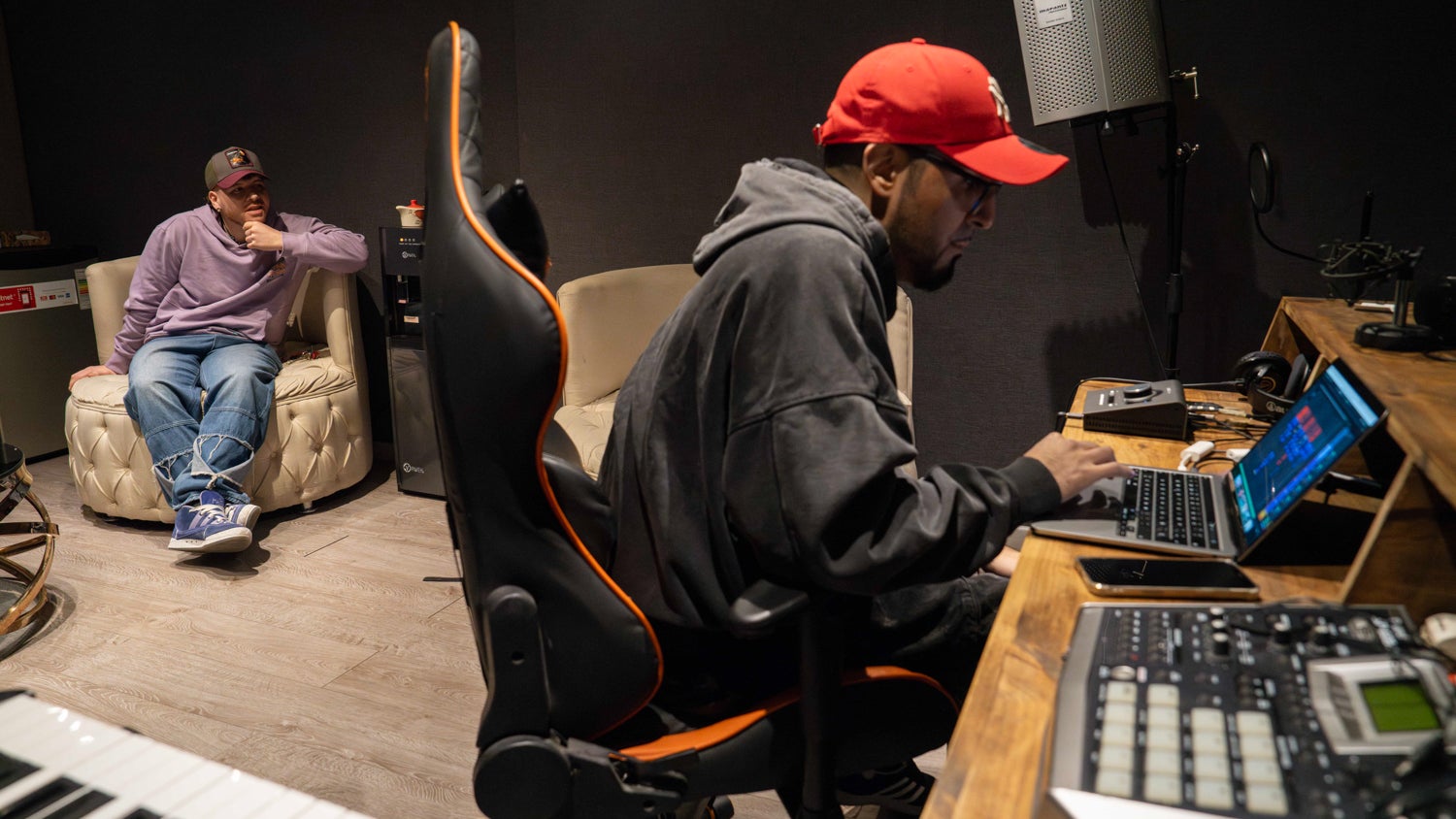A photograph of a music recording studio with Mauricio Bustos (left) seated on a chair watching Elias Adolfo Gonzales (right) using his laptop.