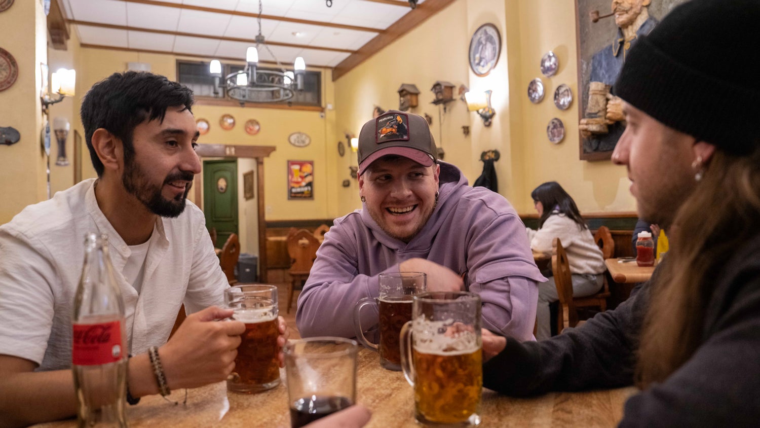 A photograph of Mauricio Bustos (center) wearing a purple hoodie and cap at a table seated between two male friends. They are engaging in conversation with multiple beer mugs in front of them.