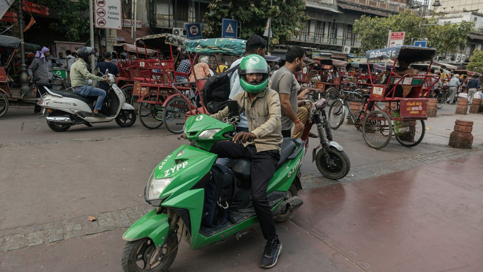 A photograph showing a man wearing a green motorcycle helmet, seating on a green scooter, in a busy urban environment.