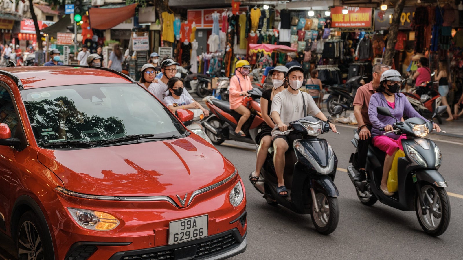 A vibrant street scene with a red VinFast car in the foreground and several motorbikes carrying riders wearing helmets and masks. The background shows shops with colorful clothing and goods, along with pedestrians waiting to cross the street.