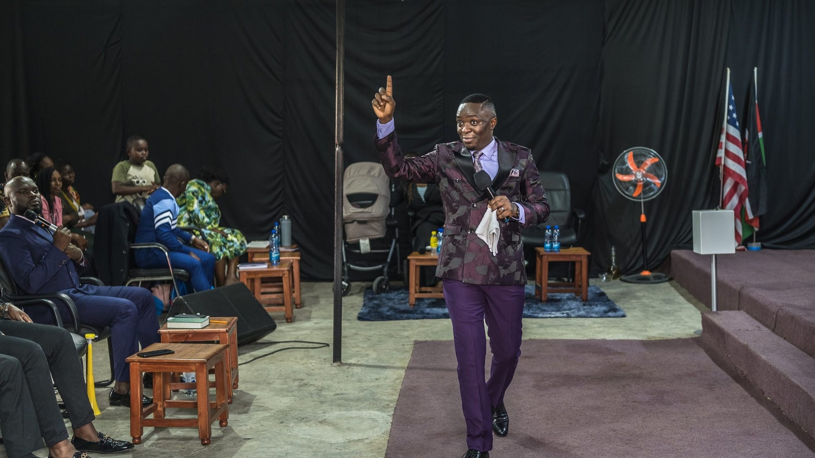 A photograph showing a man wearing purple, walking at the front of a room, with his finger raised while holding a microphone.