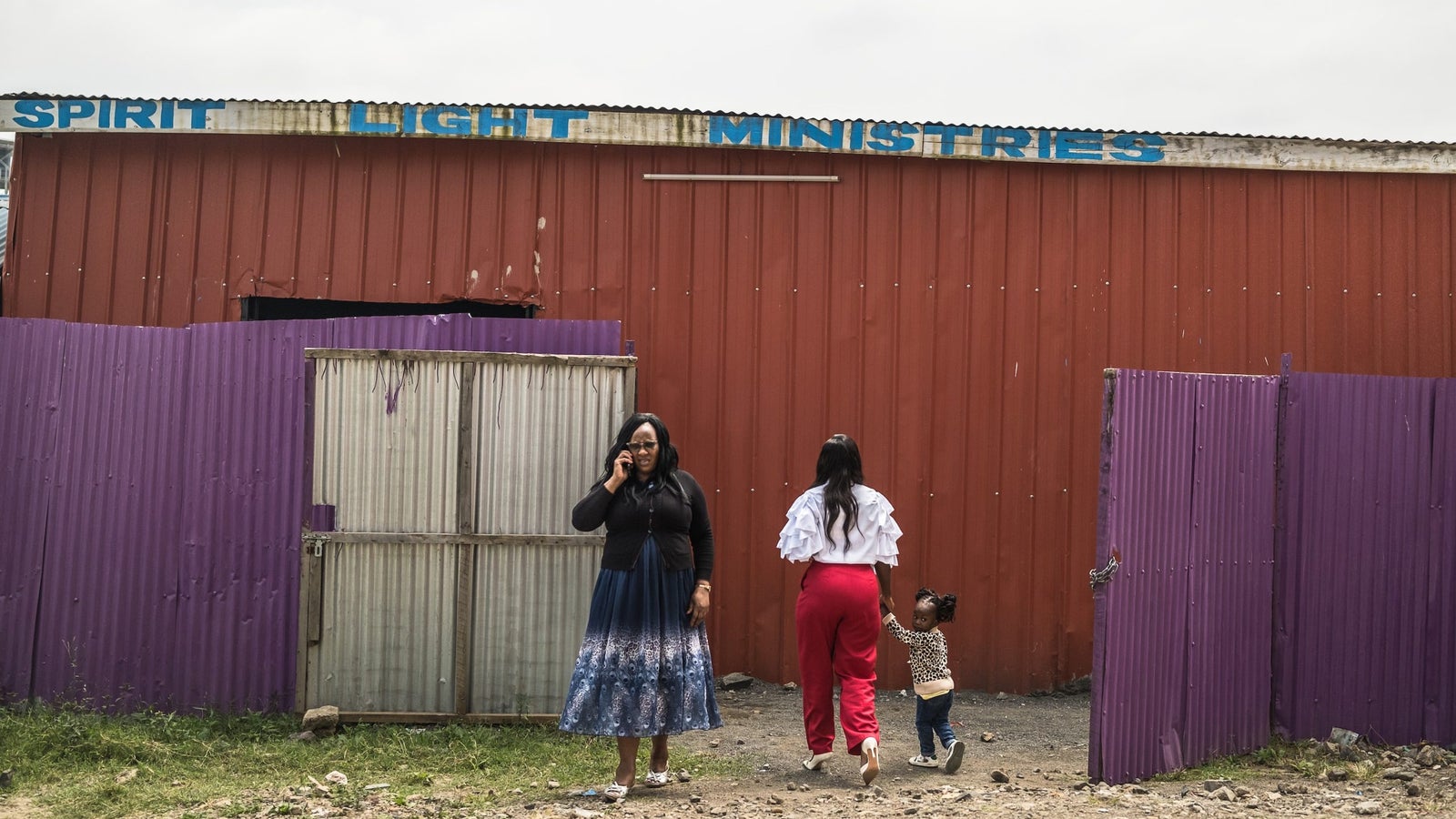 A photograph showing two women and small girl in front of a corrugated metal building and fence, with the words "Spirit Light Ministries" painting on the roof edge.