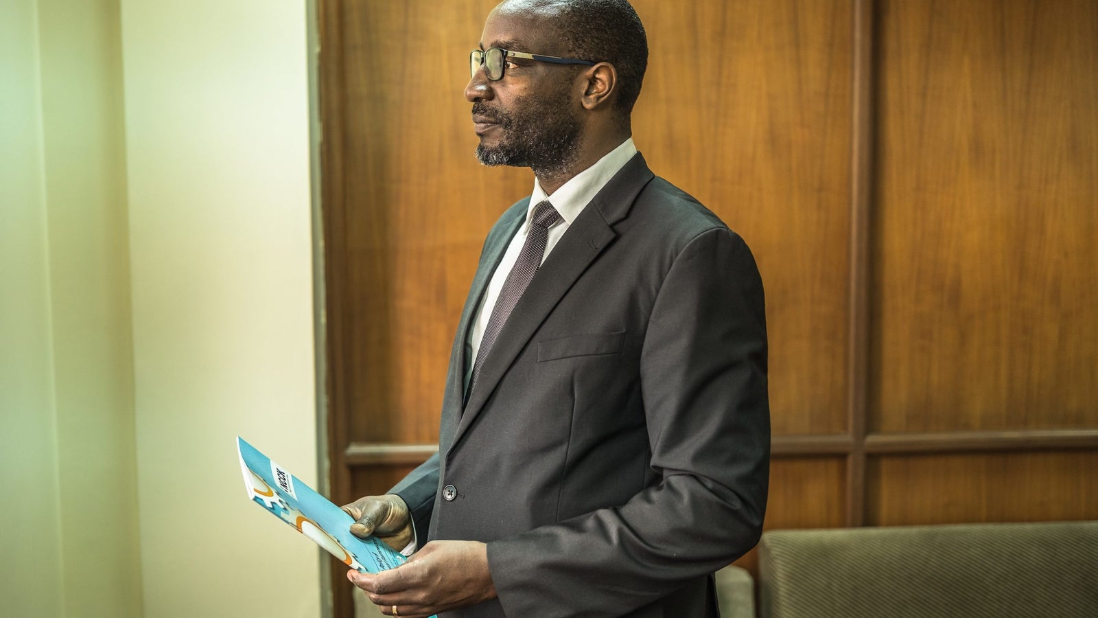 A photograph showing a portrait of a man wearing a black suit and holding a pamphlet.