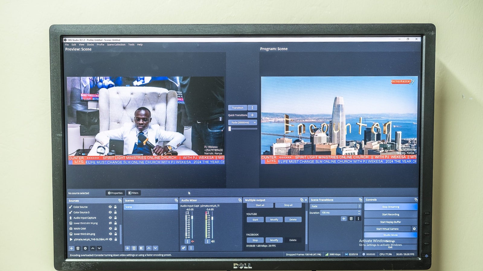 A photograph showing a television screen mounted to a wall, on the screen are two side by side programs, one with a man in white on a chair, the other of a cityscape.