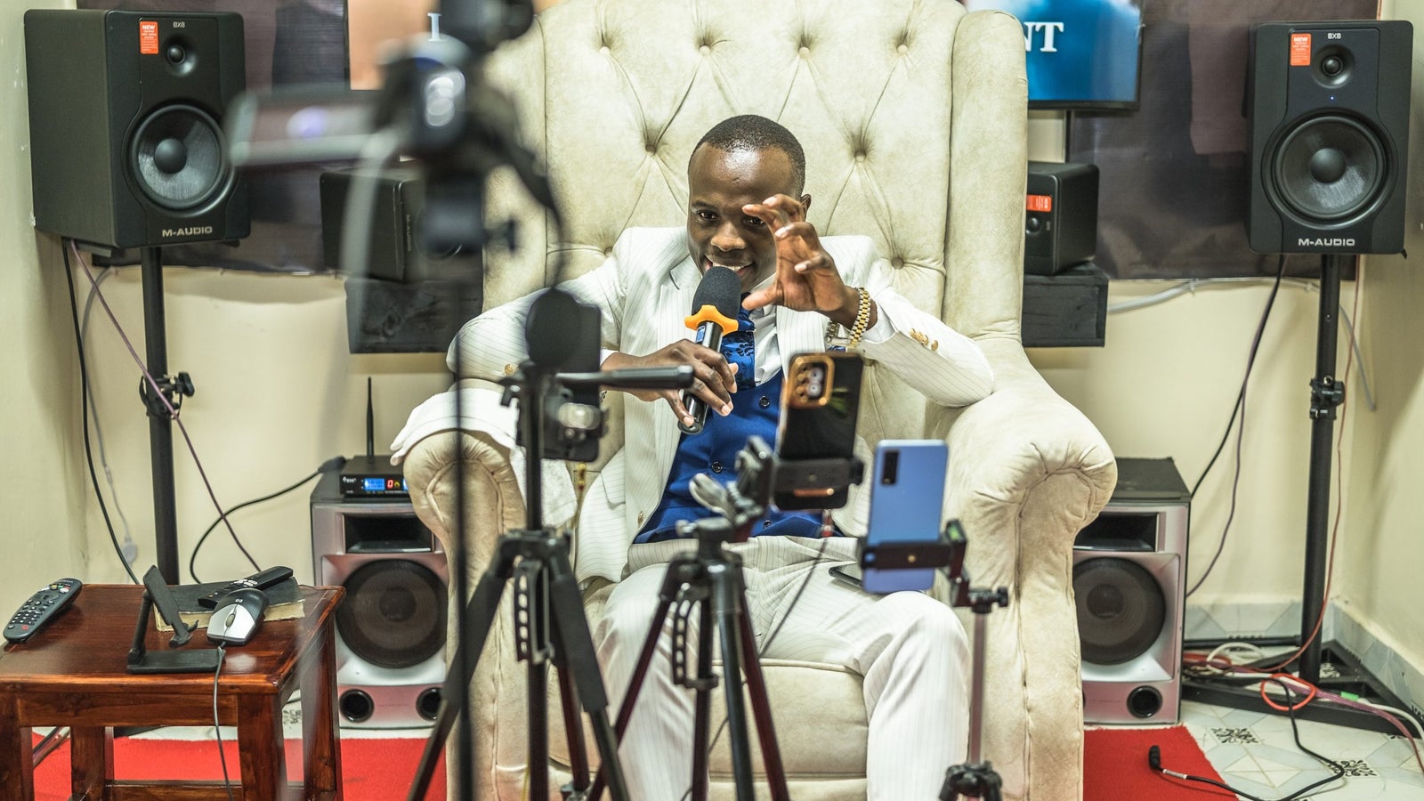 A photograph showing a man in a white suit, seated on a white chair, speaking into a microphone while staring into a number of cell phones positioned on tripods in front of him.