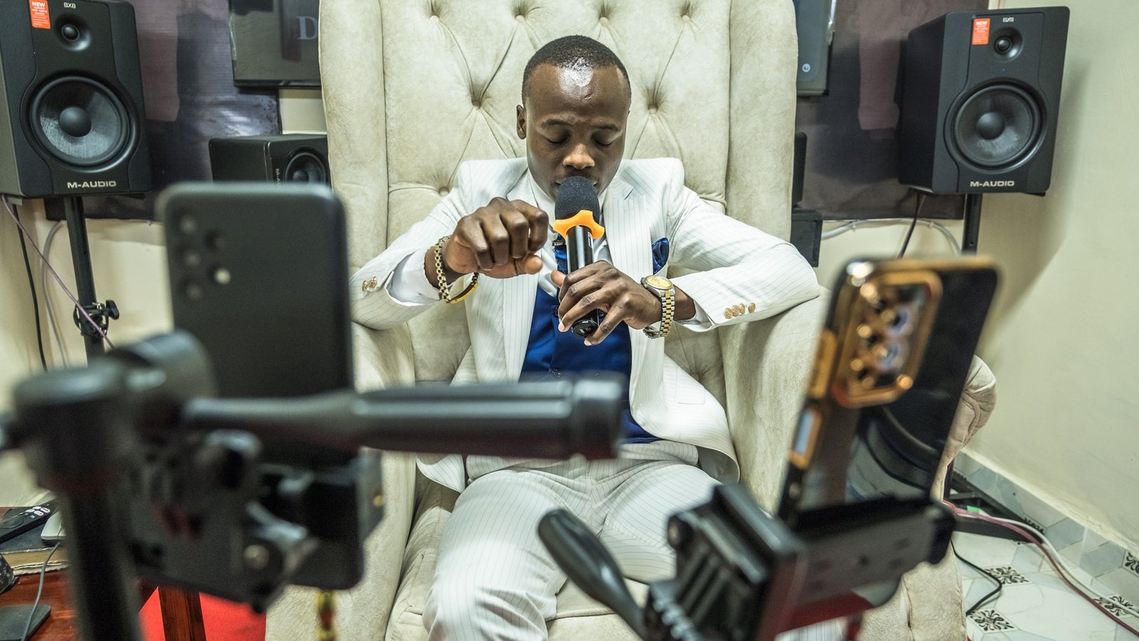 A photograph showing a man in a white suit, seated on a white chair, speaking into a microphone while staring into a number of cell phones positioned on tripods in front of him.