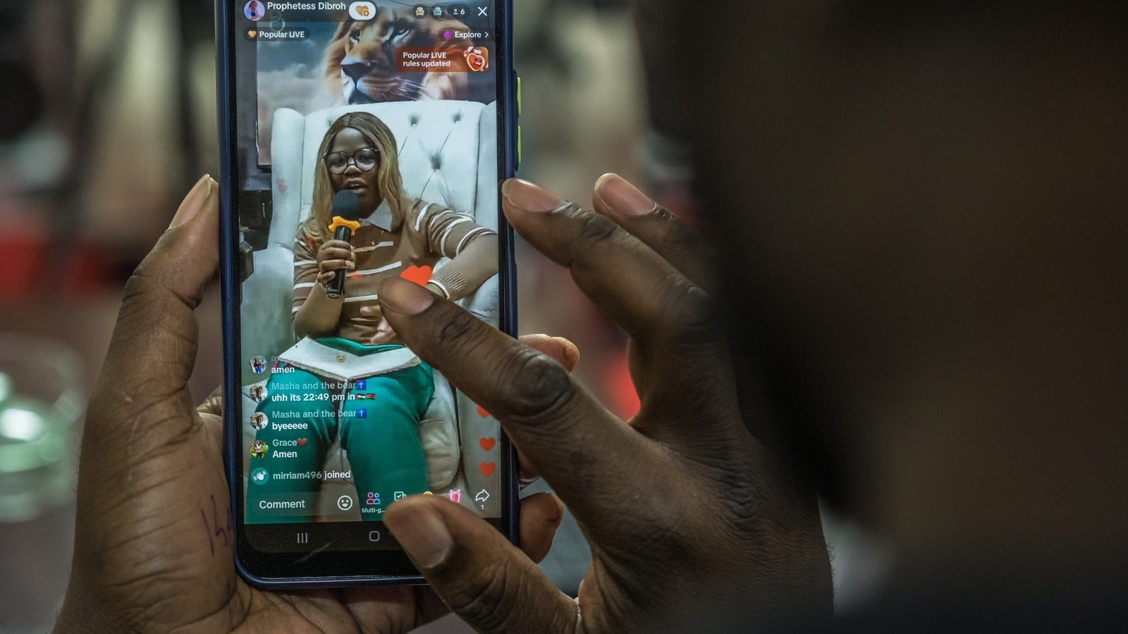A photograph showing hands using a mobile phone, on the display screen is a woman speaking into a microphone.