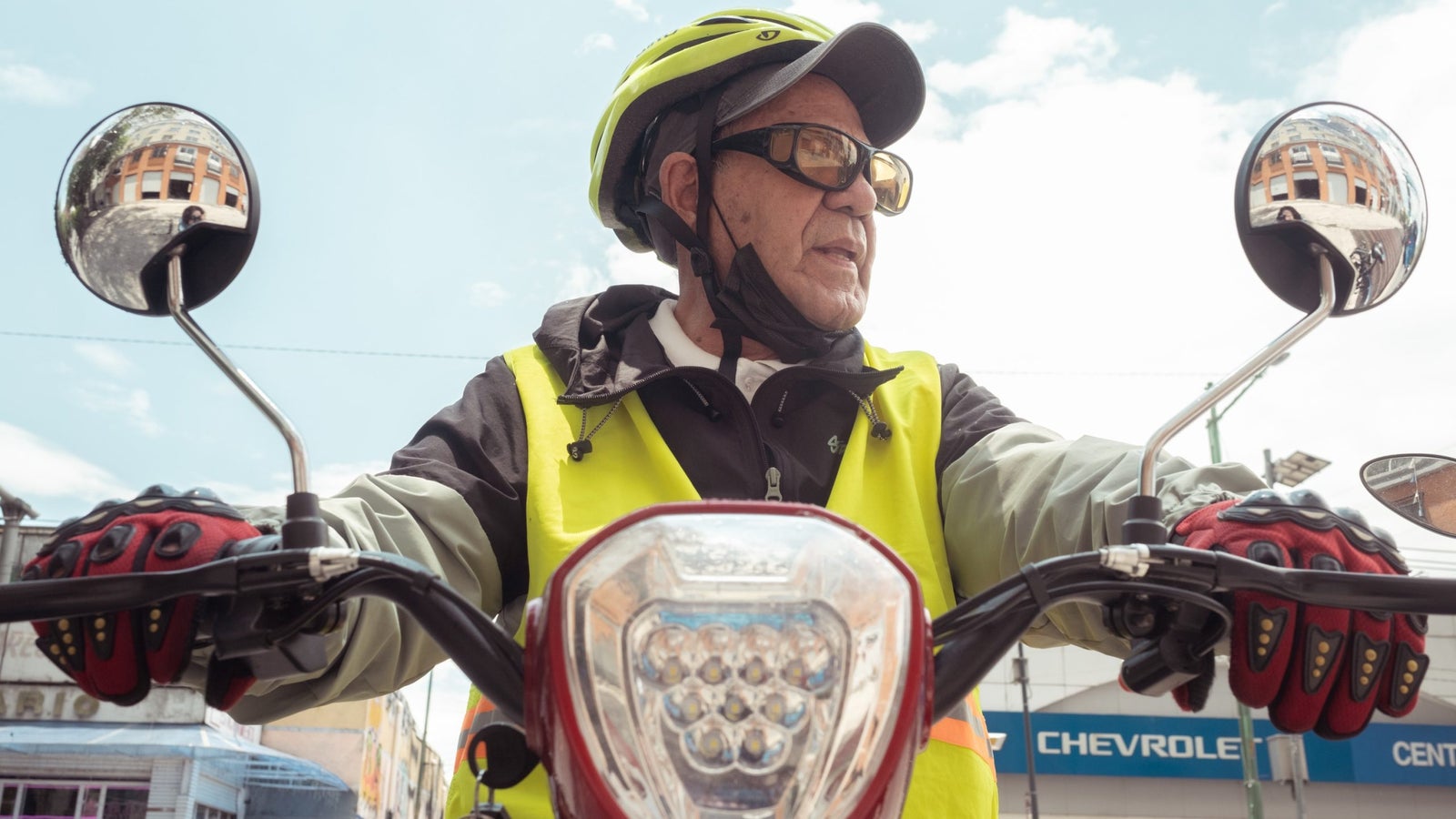 A photograph showing a man looking to the right, seated behind handlebars of a motorbike during a bright sunny day.