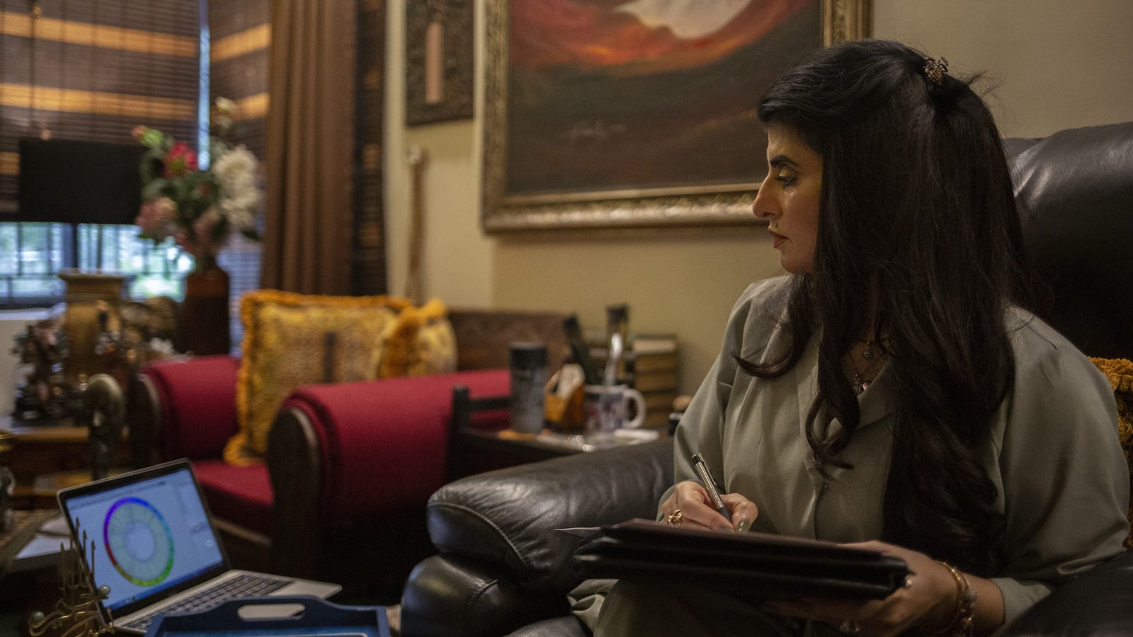 A photograph showing a woman writing on a notepad while looking at a computer screen, while seated in a large, ornate room.
