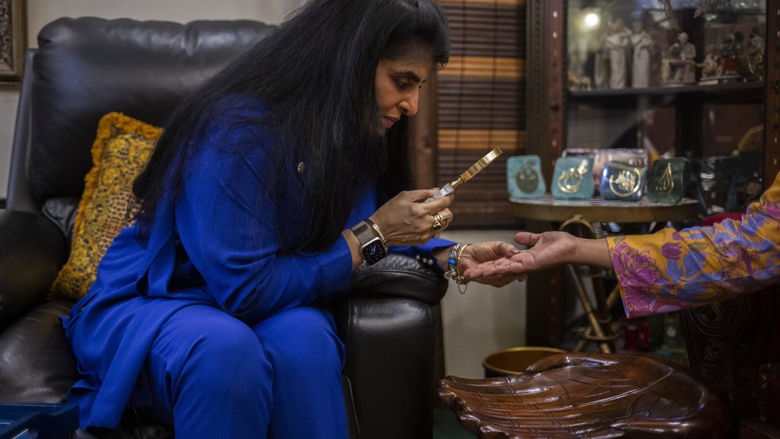 A photography of a woman with dark hair and wearing an all blue outfit, using a magnifying glass to study the hand of another person.