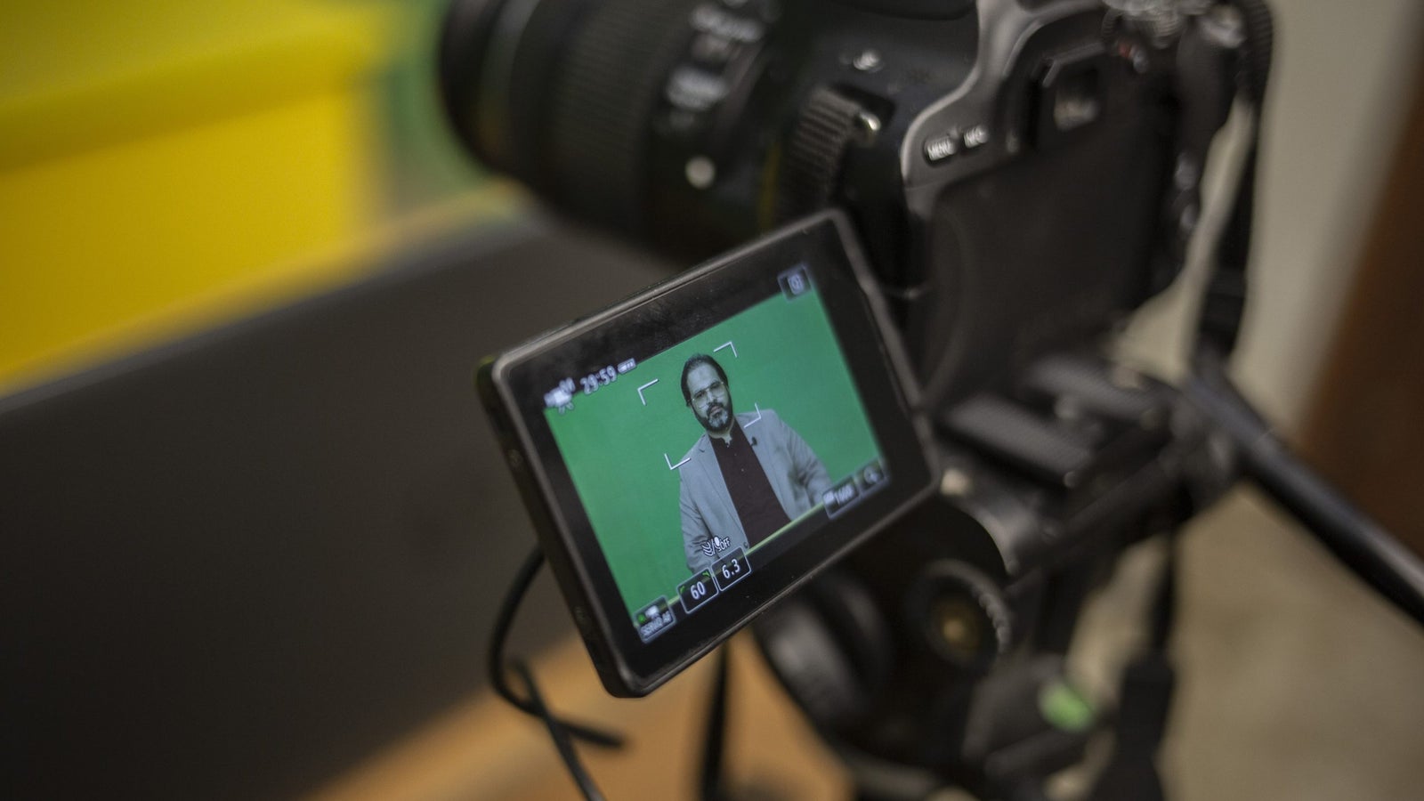 A detail photograph of a DSLR camera on a tripod and it's display screen showing the image of a man against a green screen.