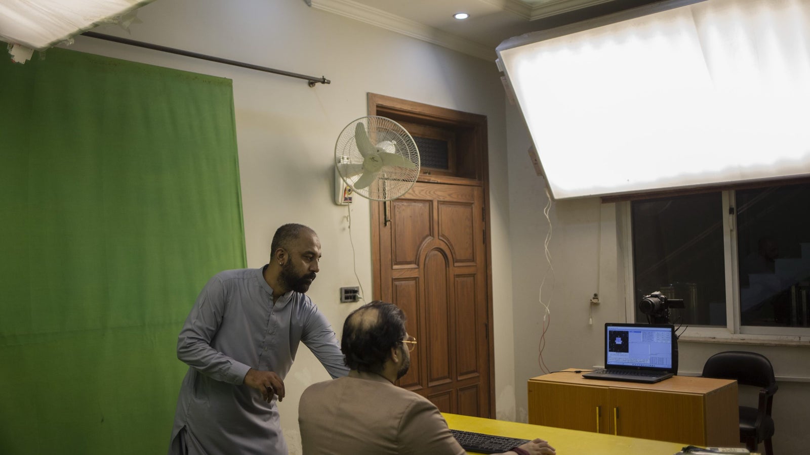 A photography of two men inside a room with large white lights, a green screen, and a computer.