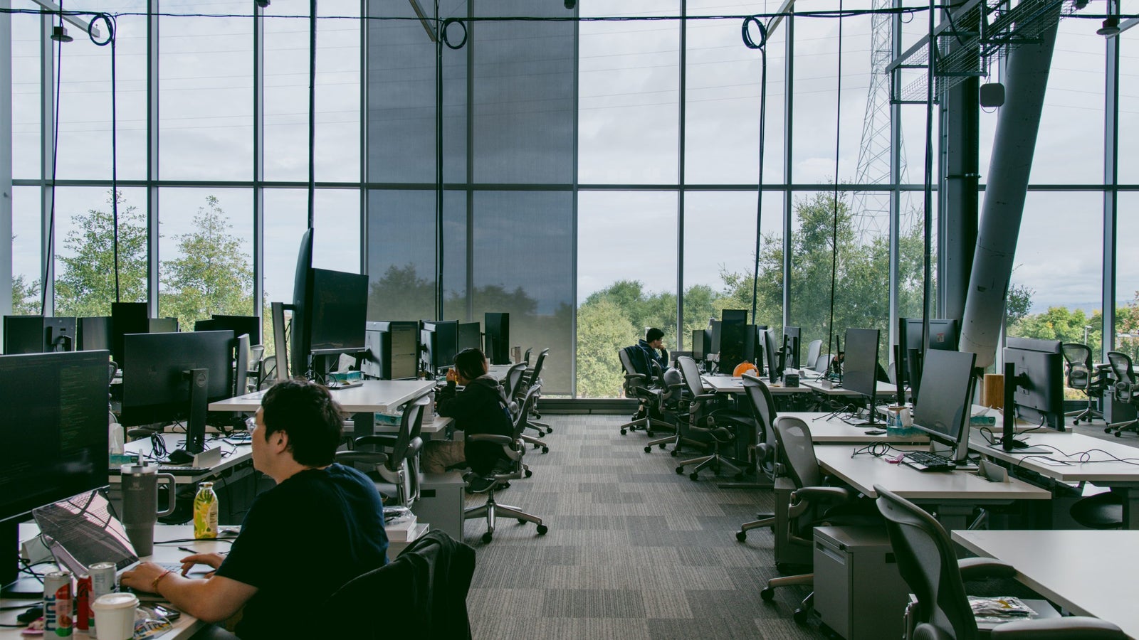 A photo of Meta’s MPK 21 corporate campus in Menlo Park showing a spacious office with large windows and several workstations equipped with monitors. People are seated at desks, engaged in work, with greenery visible outside.