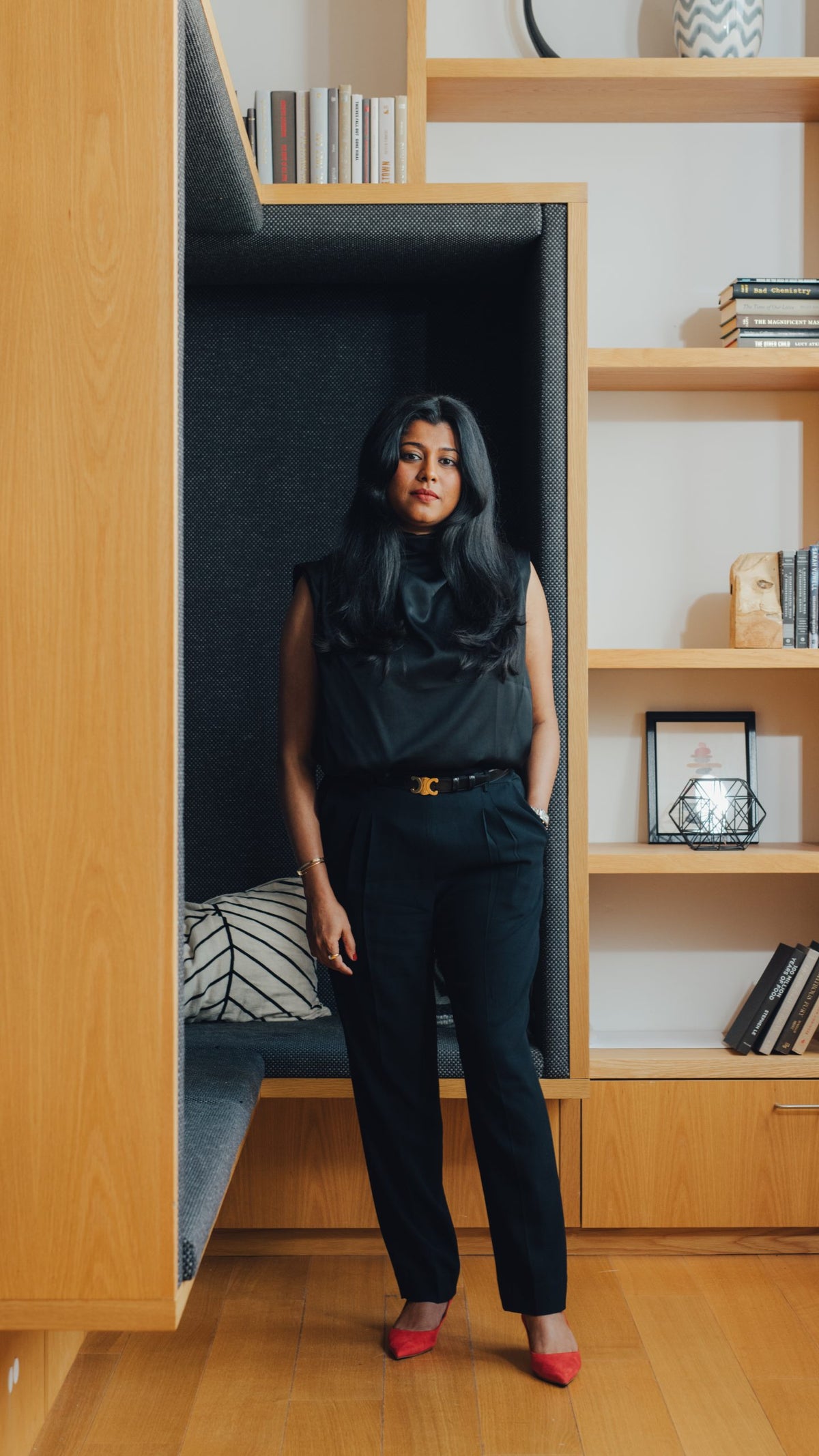 A portrait of Nikila Srinivasan at Meta's office in New York wearing a black sleeveless top and tailored pants, with red heels. She poses in a niche surrounded by wooden bookshelves filled with books and decorative items.