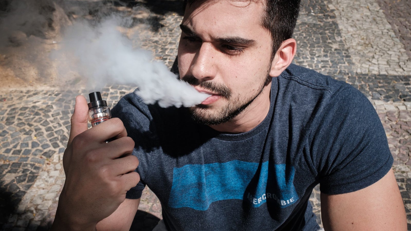 A photography showing a close image of a man blowing smoke from his mouth and clutching an e-cigarette.
