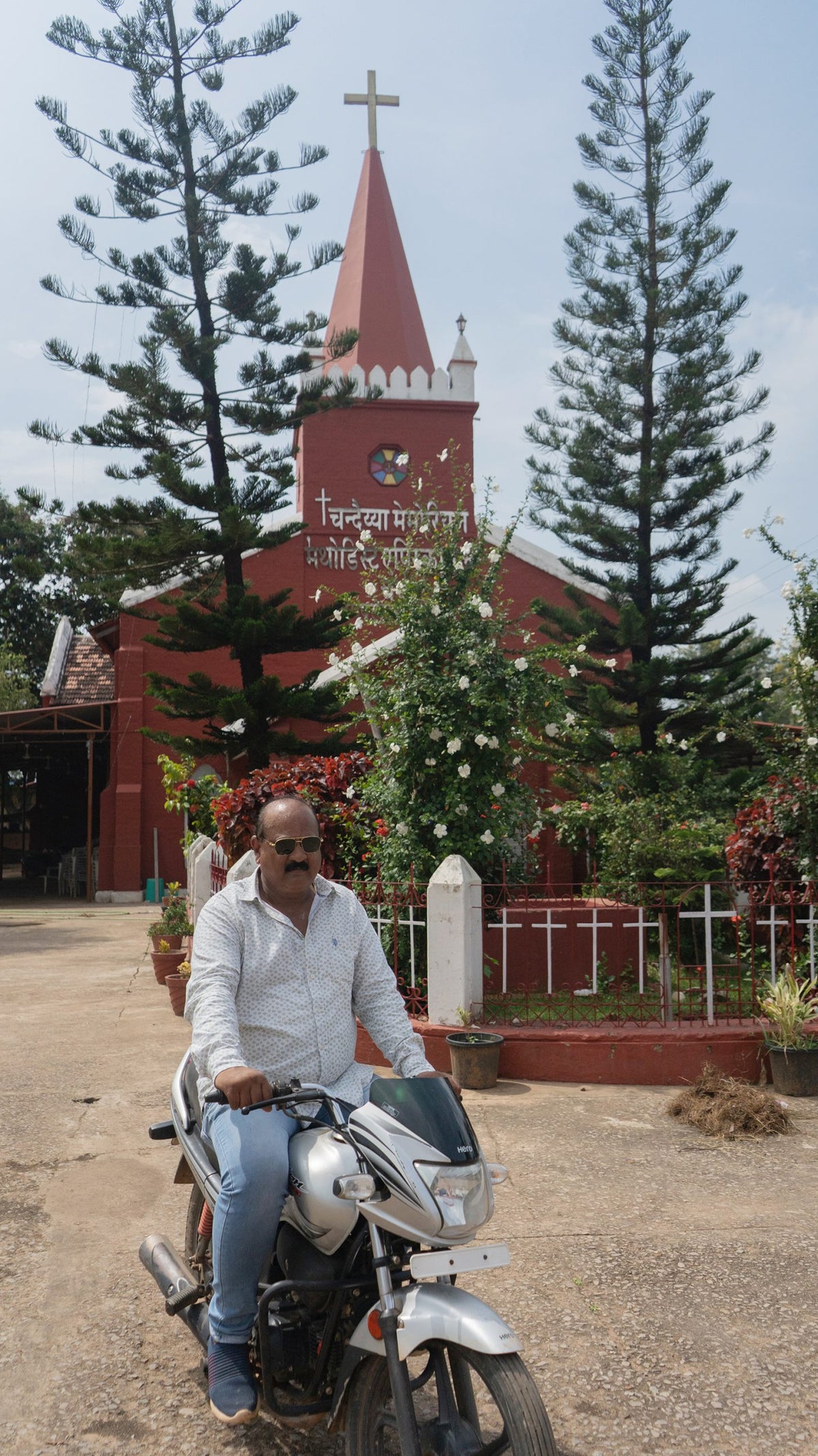 A photo of a man riding a motorcycle in front of a church with a red exterior and tall pine trees.