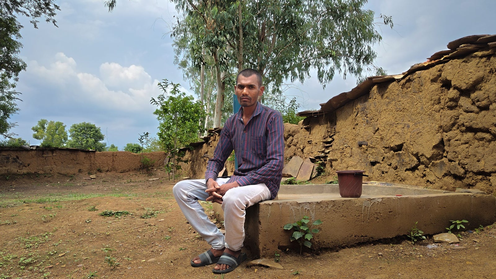 A photo of a man in a striped shirt and pants seated on the edge of a concrete structure in an open area surrounded by mud walls.