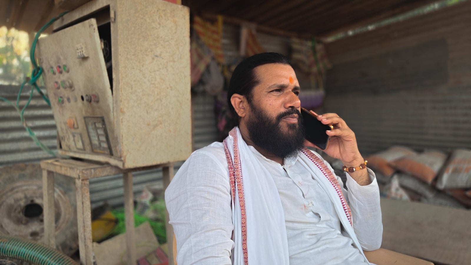 A photo of a man wearing a white Kurt and shawl with speaking on a phone. He is seated on a chair inside a structure made of roofing sheets.