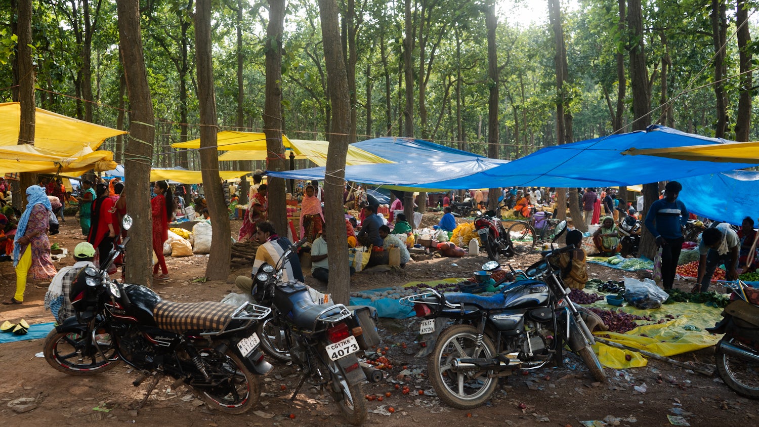 A photo of a crowded marketplace in a forest with tarp sheets tied to tree barks and motorbikes parked besides them.