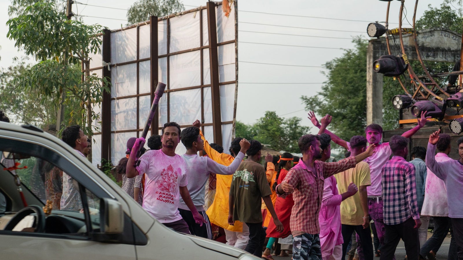 A photo of people covered in pink-colored powder celebrating on the streets. A car enters the frame from the left.