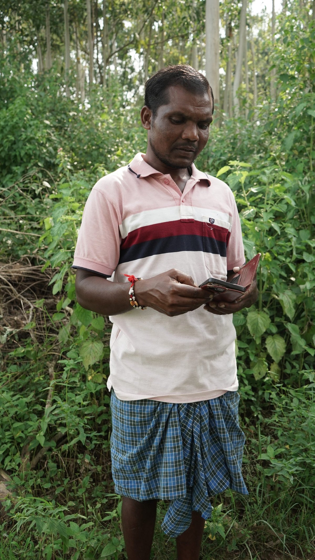 A photo of man wearing a sarong standing in front of a thick forest checking his phone.
