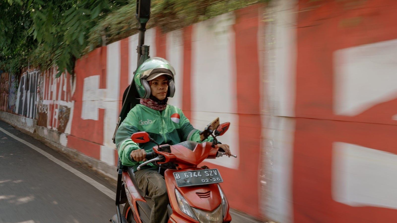 A person wearing a green Grab delivery jacket and helmet rides a red motorcycle on a narrow street, with a camera mount attached to the back of the bike.