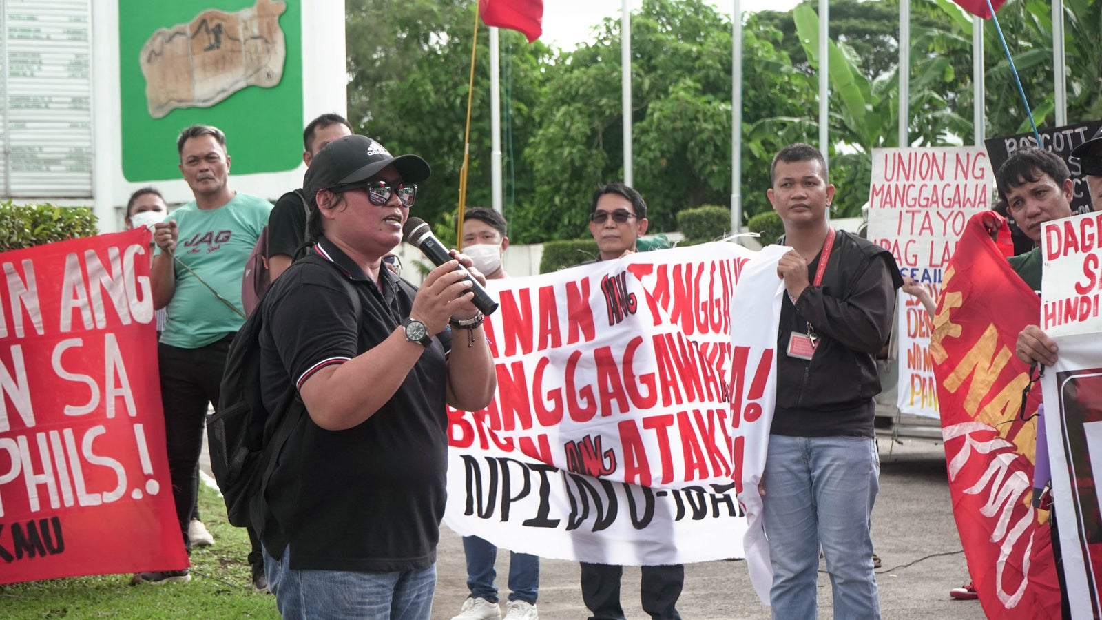 A photo of a protest showing a person speaking into a microphone with others holding banners and flags.