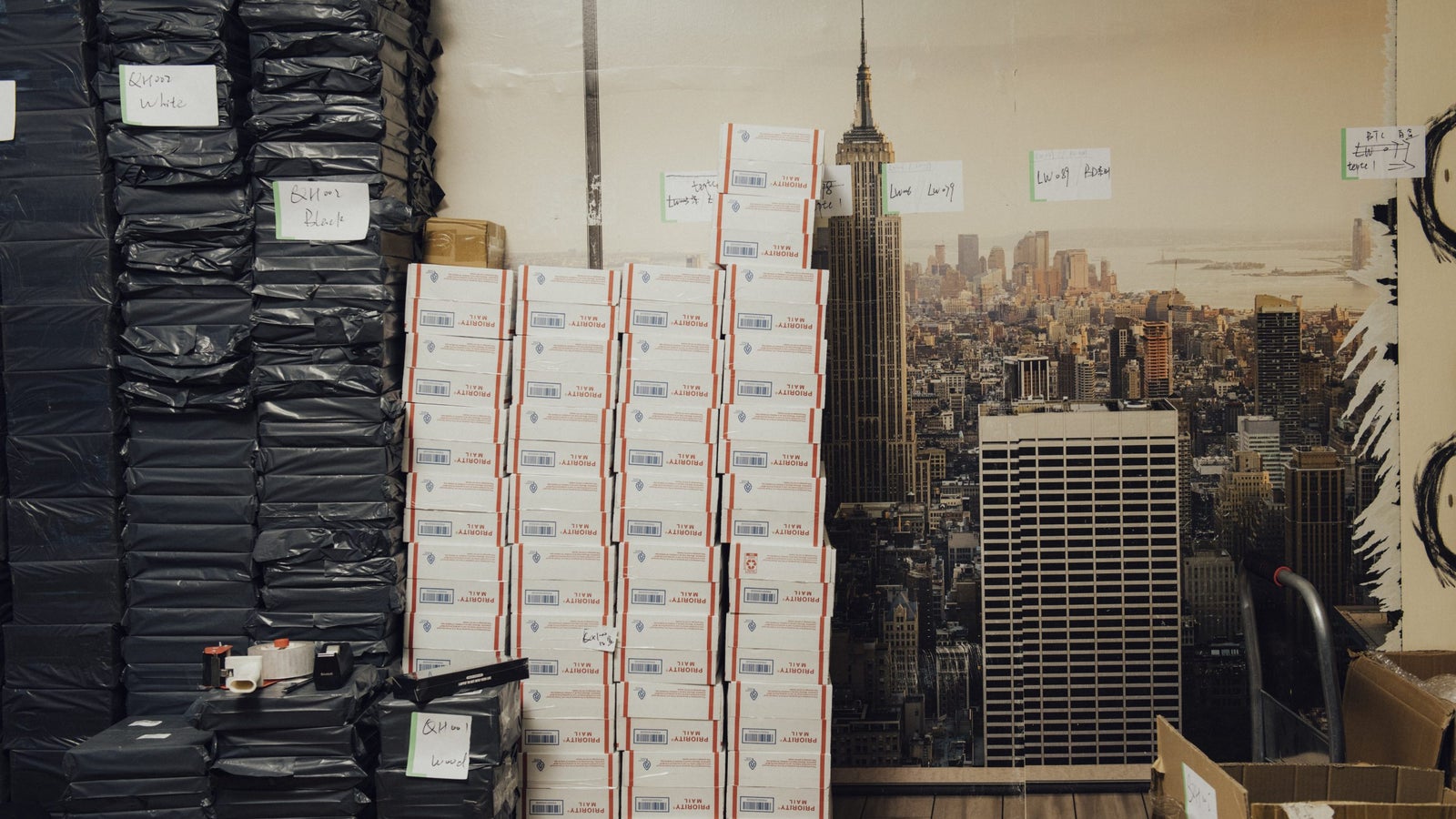A storage area with stacked black packages on the left and a tall stack of white and red mailing boxes on the right. In the background, there is a wallpaper depicting a city skyline featuring the Empire State Building. Some handwritten labels are attached to the walls and boxes.
