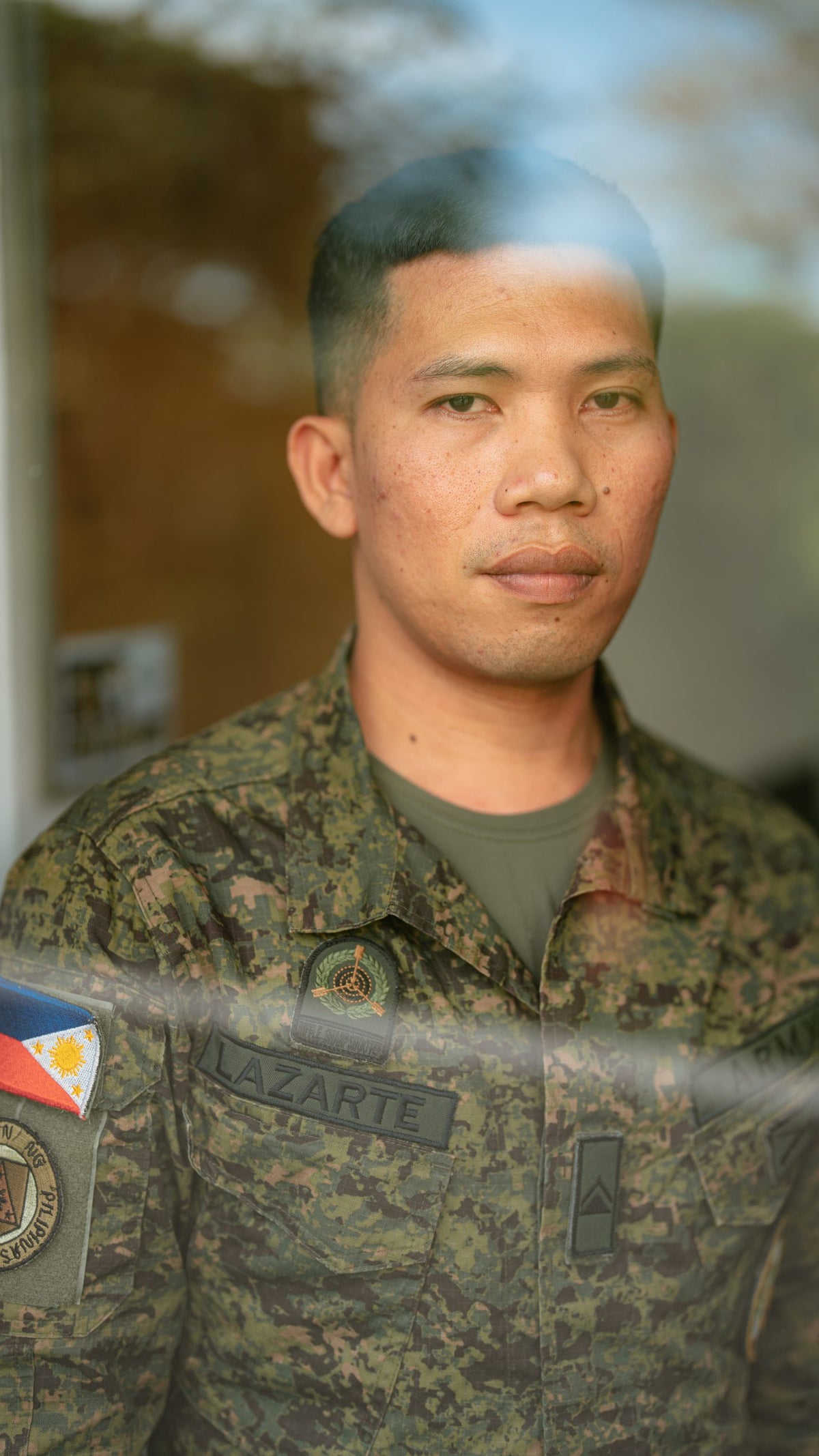 A portrait of Ferdinand Lazarte, an instructor of the Philippine Army’s Cyber Battalion, wearing military uniform and taken through a glass, which reflects the sky and trees outside.