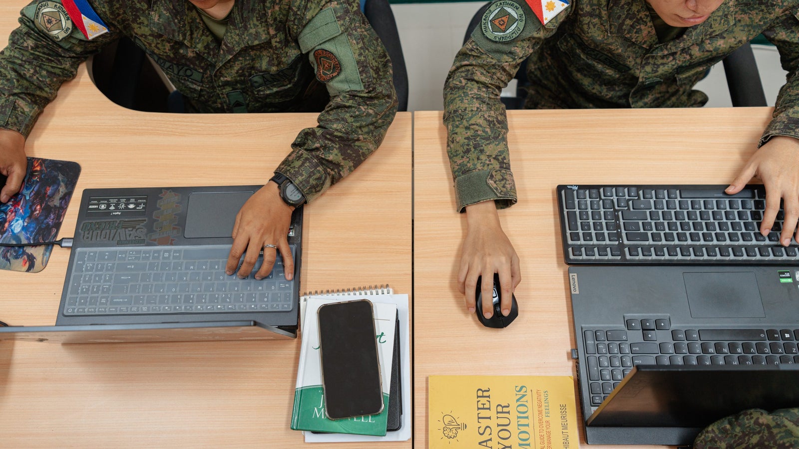 Two military personnel are seated at a wooden table, working on laptops. One person uses a touchpad while the other operates a mouse. Various items, including notebooks and a book titled "Master Your Emotions," are visible on the table. Both individuals wear military uniforms that display patches, including flags.