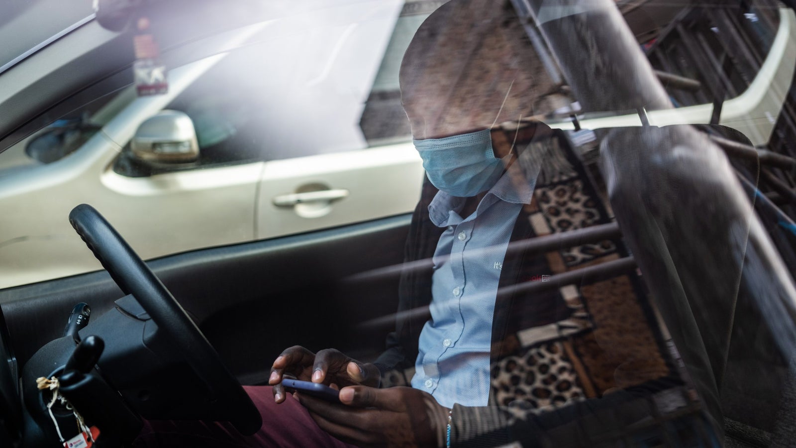 A photograph taken through a car window of a man seated in the driver's seat using his phone, wearing a face mask. The glass window shows a reflection of the streets.