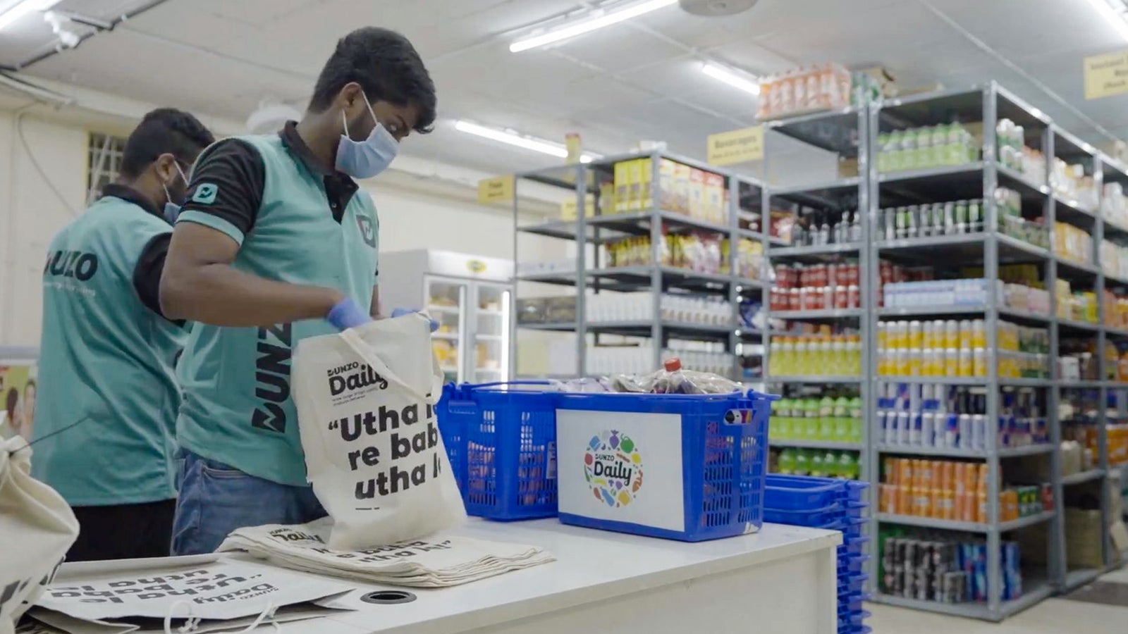 Two workers wearing masks and Dunzo uniforms are arranging items in a dark store. One is packing a cloth bag with groceries while the other is organizing baskets filled with products on a table, surrounded by shelves stocked with various goods.