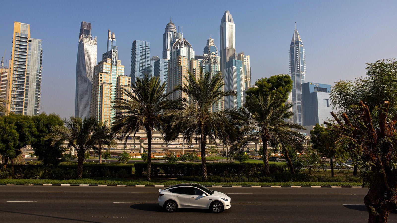 A white car drives along a road lined with palm trees, with a striking skyline of modern skyscrapers in the background under a clear blue sky.