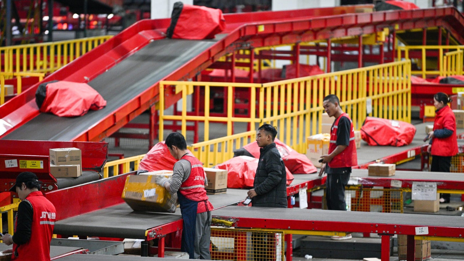 Workers in red vests sorting packages on conveyor belts in a large warehouse, with parcels and bags on the conveyor and the floor.